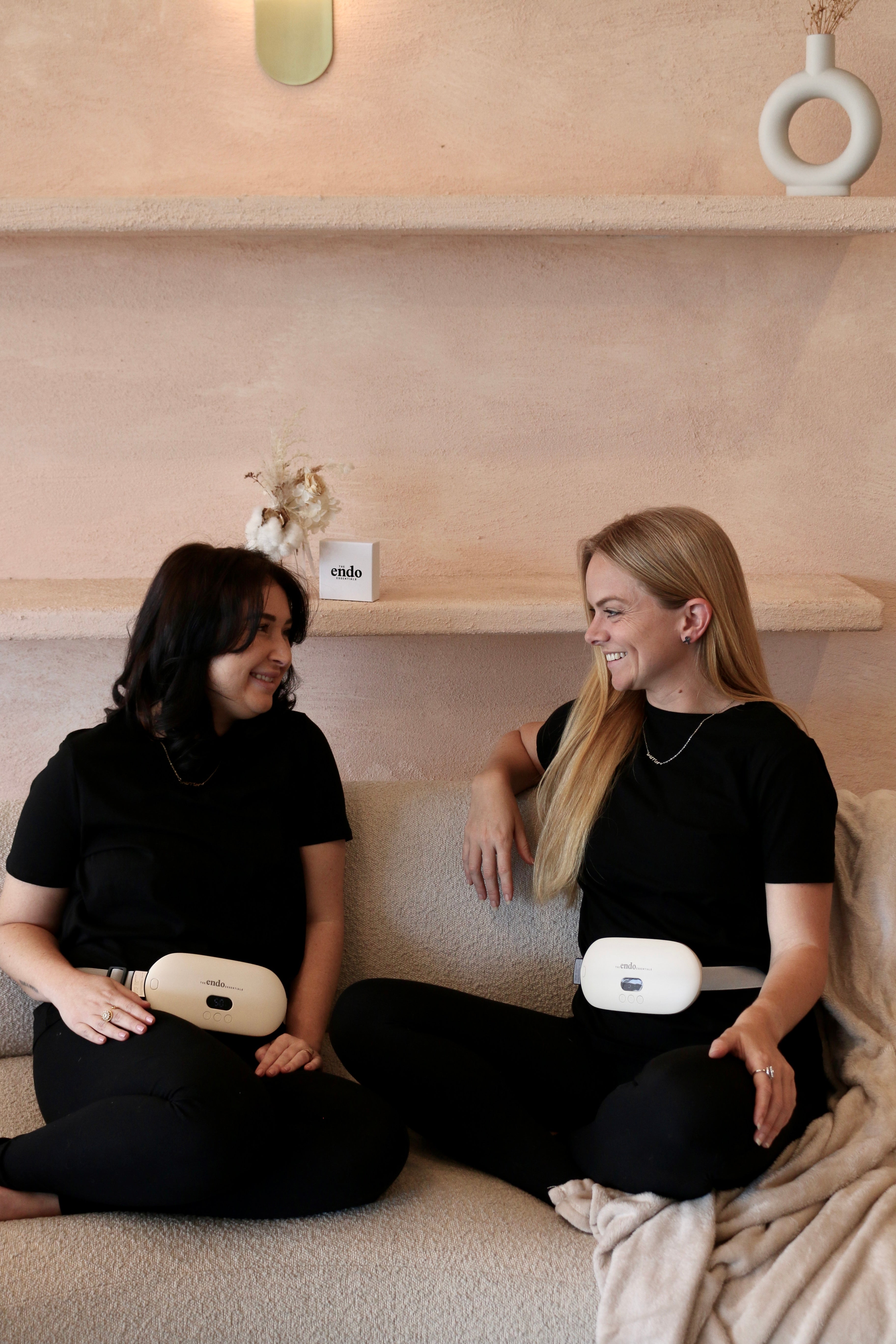 Two women sitting on a couch wearing black outfits with portable Electric Heating Belts, smiling and looking at each other.