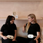 Two women sitting on a couch wearing black outfits with portable Electric Heating Belts, smiling and looking at each other.