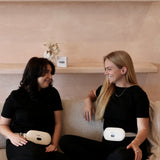 Two women sitting on a couch wearing black outfits with portable Electric Heating Belts, smiling and looking at each other.