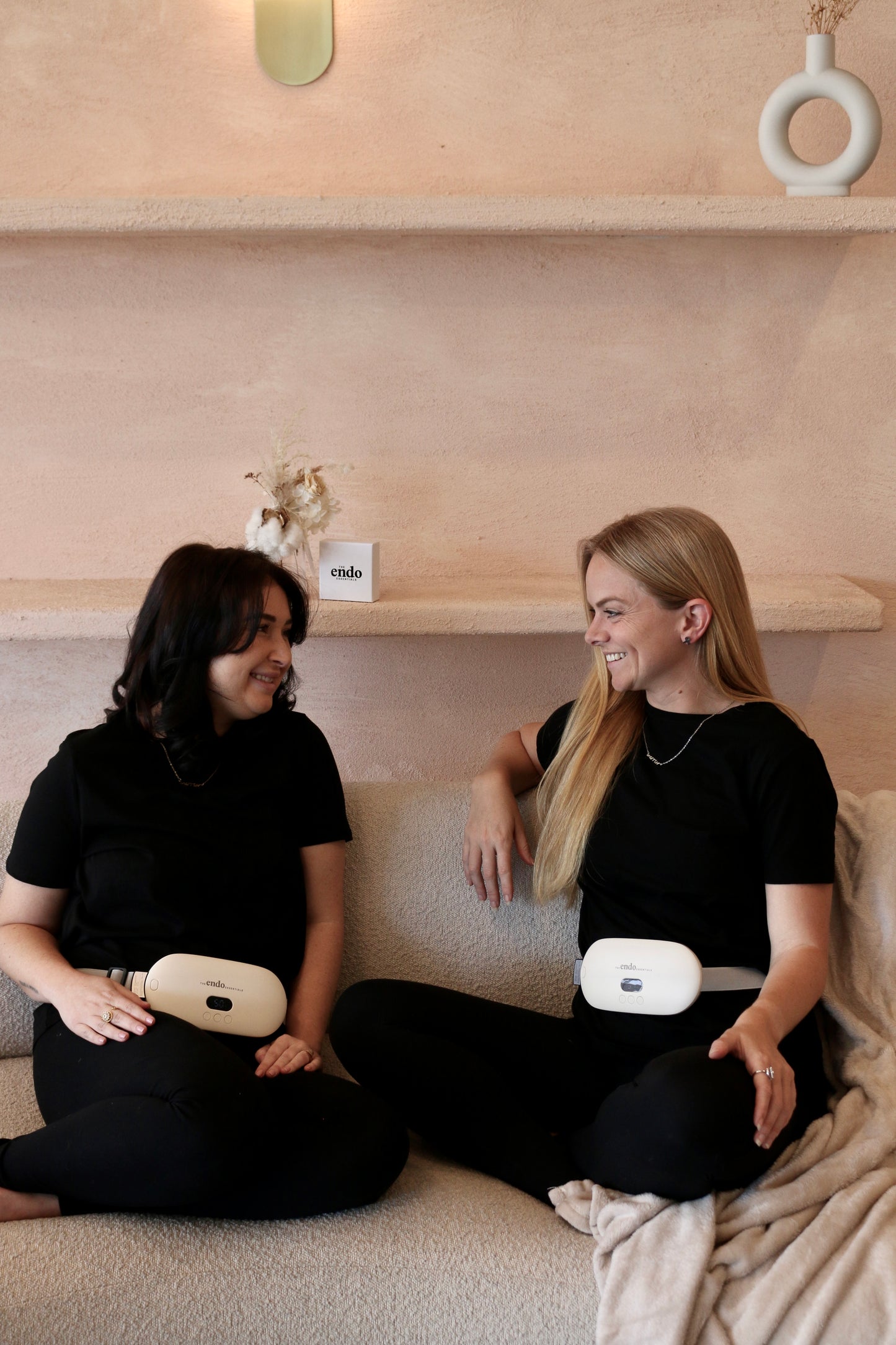 Two women sitting on a couch wearing black outfits with portable Electric Heating Belts, smiling and looking at each other.
