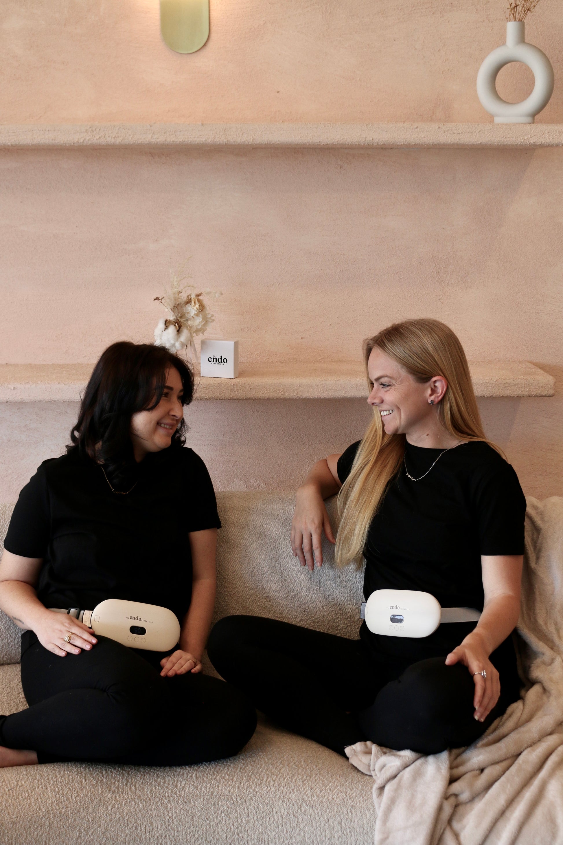 Two women sitting on a couch wearing black outfits with portable Electric Heating Belts, smiling and looking at each other.